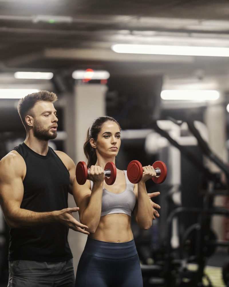 A fit sportswoman in shape is doing exercises with dumbbells in a gym and her coach is helping her.