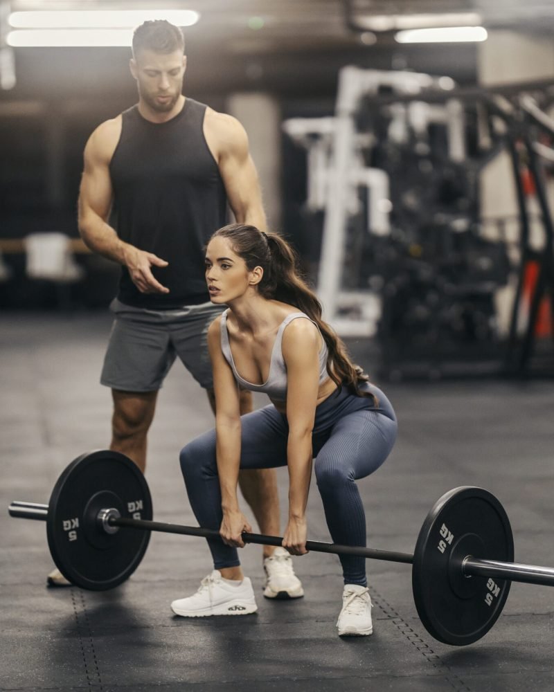A strong sportswoman is lifting weights in a gym while her instructor is helping her.